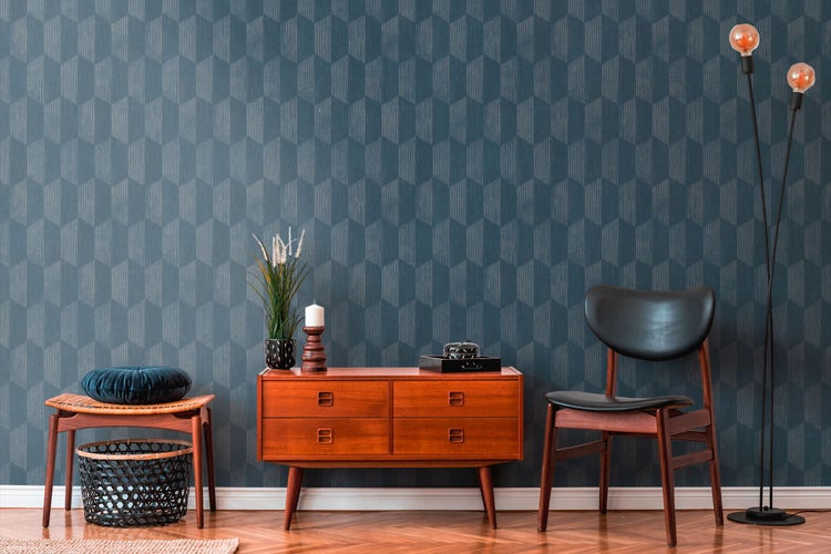 Wohnzimmer mit Kommode, Stuhl, Hocker und Stehlampe vor einer Wand mit geometrischer Tapete.