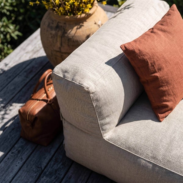 Outdoor Lounge Sofa mit strukturiertem Stoffbezug, einem terrakottafarbenen Kissen und einer braunen Ledertasche auf einer Holzterrasse.