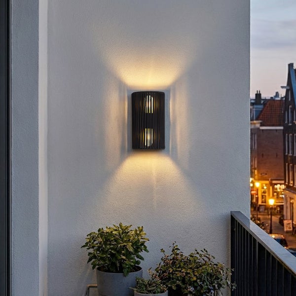 Moderne Außenwandleuchte mit zwei Lichtkegeln montiert an einer hellen Hauswand auf einem Balkon mit Topfpflanzen bei Abenddämmerung.