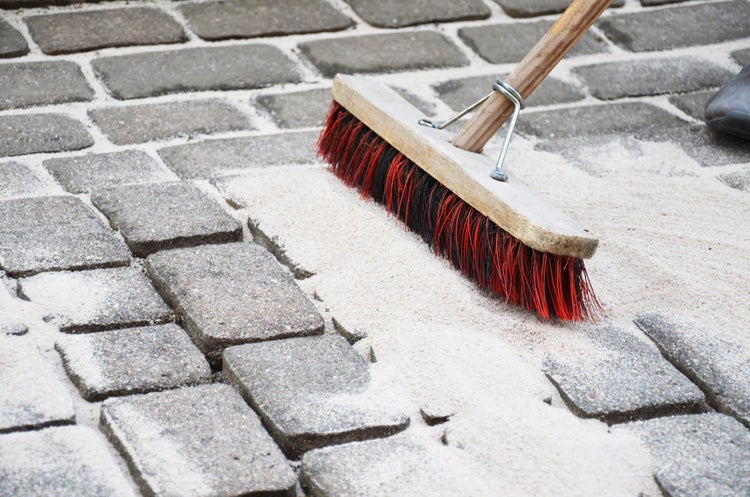 Ein Besen fegt Fugensand in die Zwischenräume von Kopfsteinpflaster, um die Pflastersteine auf einer Terrasse oder einem Weg zu stabilisieren.