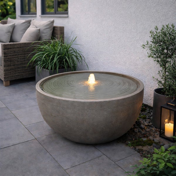 Moderner runder Gartenbrunnen in Steinoptik mit beleuchtetem Wasserspiel auf einer Terrasse neben Loungemöbeln und Pflanzen.