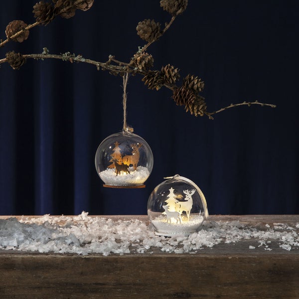 Dekorative Glaskugeln mit Tierfiguren und Beleuchtung auf einem Holztisch mit Kunstschnee und einem Ast mit Tannenzapfen