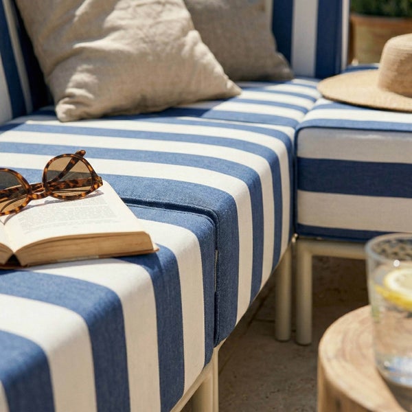 Blau-weiß gestreifte Gartenlounge mit Polsterauflage, Kissen, einem Buch und einer Sonnenbrille auf der Sitzfläche.