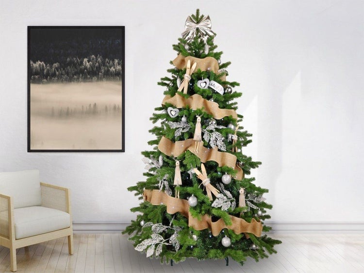 Geschmückter Weihnachtsbaum mit Juteband, Holzschmuck, Skiern und silberfarbenen Dekoelementen in einem Wohnraum mit Sessel und Wandbild.