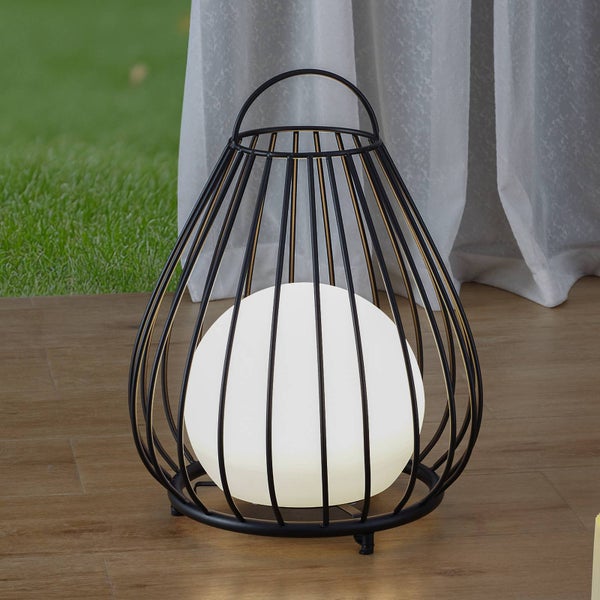 Moderne Käfiglaterne aus schwarzem Metall mit weißer Kugelleuchte auf einer Holzterrasse vor Gras und hellem Vorhang.
