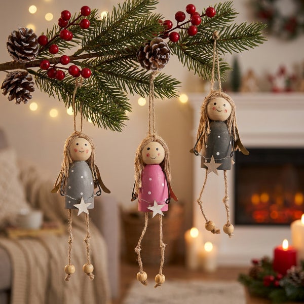 Drei hängende Weihnachtsengel aus Holz als Baumschmuck, hängend an einem Tannenzweig mit Beeren und Zapfen vor einem Kamin.
