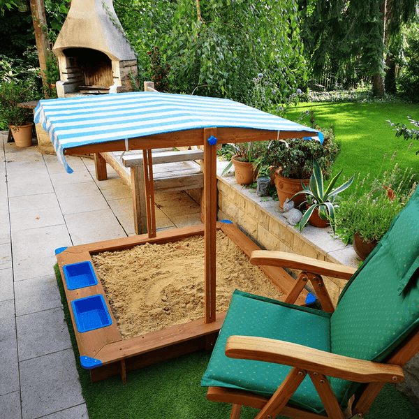 Kinder-Sandkasten aus Holz mit gestreiftem Dach und blauen Kunststoffeinsätzen auf einer Terrasse im Garten.