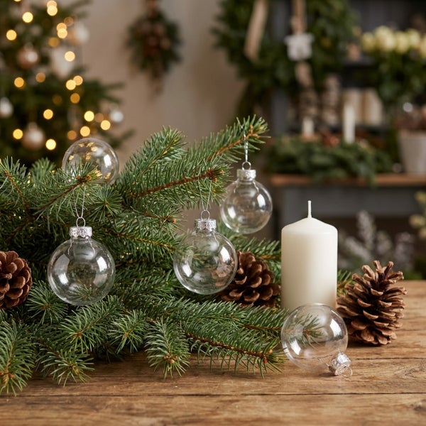 Weihnachtliche Tischdekoration mit Tannenzweig, klaren Glaskugeln, Zapfen und einer Kerze auf rustikalem Holztisch.