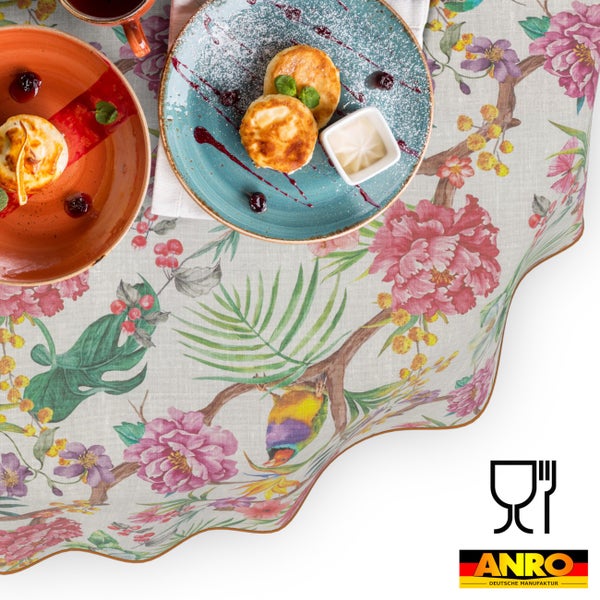 Tischdecke mit Blumenmuster und Vogelmotiv, angerichtet mit Speisetellern. Wellenrand. Symbol für Lebensmittelechtheit und ANRO DEUTSCHE MANUFAKTUR Logo.
