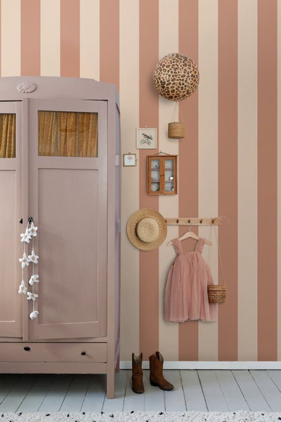 Vliestapete mit vertikalen Streifen in Terrakotta und Beige in einem hellen Kinderzimmer mit rosa Schrank und Boho-Dekoration.