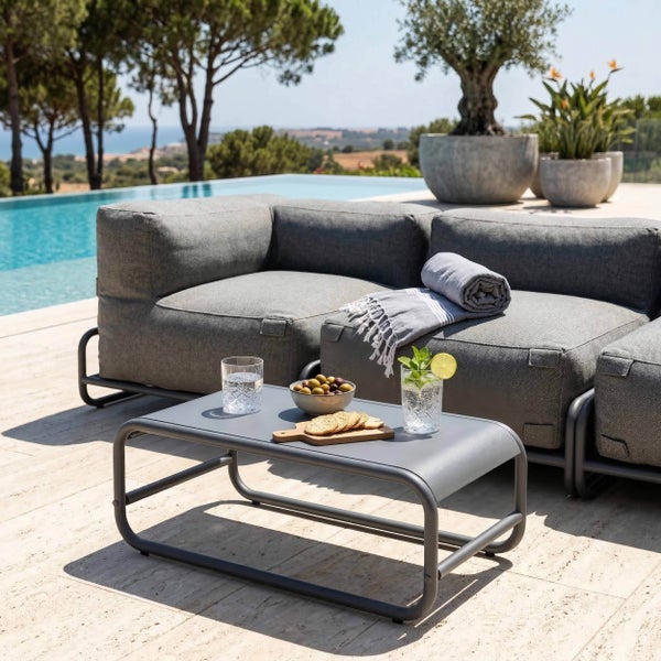 Moderne Lounge-Garnitur mit grauem Sofa und niedrigem Metalltisch auf einer hellen Terrasse neben einem Swimmingpool.