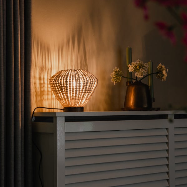 Tischlampe mit Lampenschirm aus Holzperlen auf einer weißen Heizungsverkleidung neben einer Vase mit Blumen und Kerzen in einem Wohnraum.