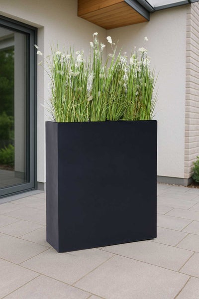 Hoher, rechteckiger Pflanzkübel in Schwarz, bepflanzt mit hohem Gras und weißen Blüten, auf einer gepflasterten Terrasse.