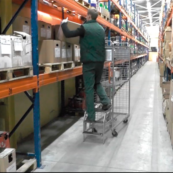 Ein Mitarbeiter in einem Lagerhaus platziert eine Kiste von einem Regal auf einen Rollwagen mit integrierter Leiter.