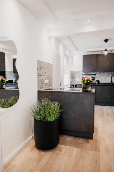 Moderne Küche mit dunkelgrauen Schränken, gemustertem Fliesenspiegel, Boden in Holzoptik, großem schwarzem Pflanztopf und gewelltem Wandspiegel.