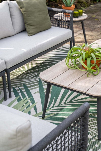 Moderne Gartenlounge mit grauem Sofa, Couchtisch in Holzoptik und grünem Outdoorteppich mit Blattmuster auf einer Terrasse.