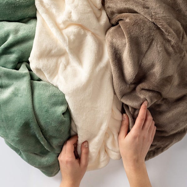 Weiche Fleece-Wohndecken in Grün, Cremeweiß und Braun werden von Händen gehalten, um Material und Struktur zu zeigen.