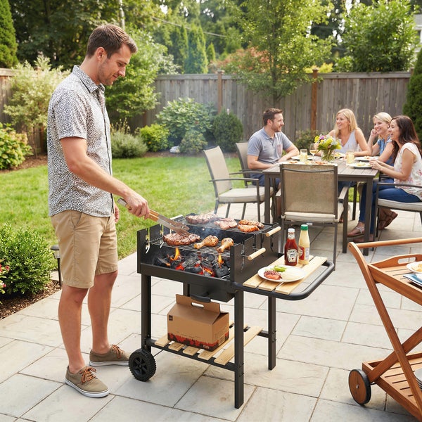 Mann grillt Fleisch auf einem Holzkohlegrill aus Metall und Holz in einem hellen Garten mit Gästen an einem Esstisch im Hintergrund.