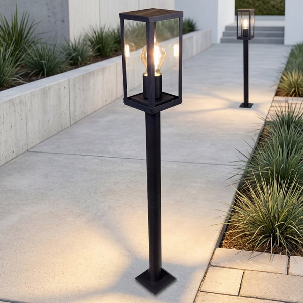 Moderne Außenstandleuchten mit rechteckigen Glaslaternen und warmweißen Filament-Leuchtmitteln an einem Gartenweg aus Beton.