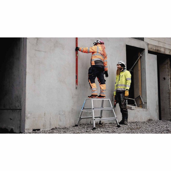 Zwei Arbeiter auf einer Baustelle, eine Person steht auf einer Arbeitsbühne und richtet eine Wasserwaage an einer Betonwand aus, die andere Person steht daneben mit einem Akkuschrauber.