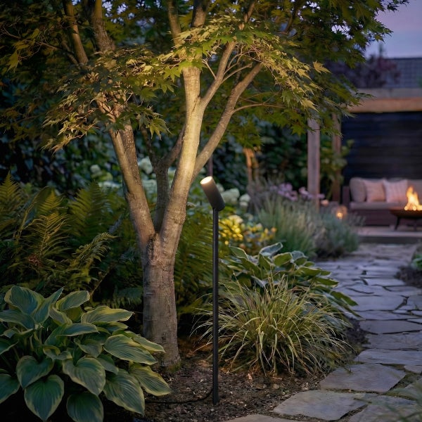 Garten-Spot auf Erdspieß beleuchtet einen Baumstamm in einem bepflanzten Garten mit Steinweg und Feuerstelle im Hintergrund.