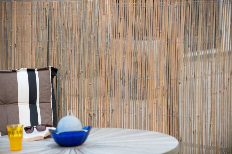 Gartenszene mit Bambuszaun, Gartentisch, Stuhlkissen, Buch, Sonnenbrille und Getränk