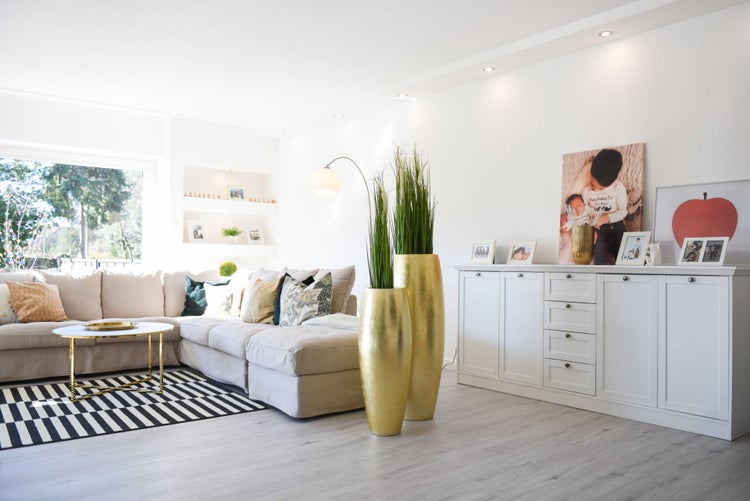 Hell eingerichtetes Wohnzimmer mit beigem Ecksofa, gestreiftem Teppich, goldfarbenen Bodenvasen und weißem Sideboard auf hellem Laminatboden.
