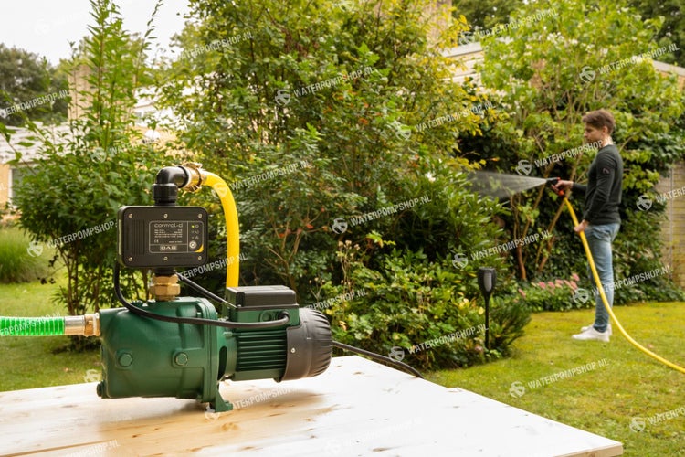 Garten mit Hauswasserwerk und Person die mit einem gelben Wasserschlauch gießt.