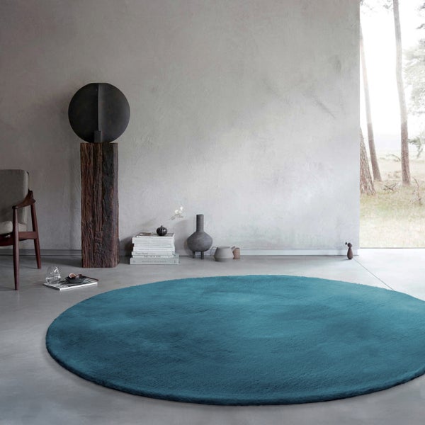 Runder Hochflor-Teppich in modernem Zimmer mit Wand in Betonoptik, Holzskulptur und Fensterausblick in einen Wald.