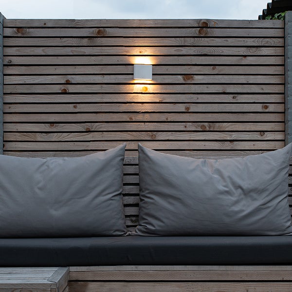Moderne Außenwandleuchte mit Lichtaustritt nach oben und unten an einer Holzwand über einer Sitzbank mit grauen Kissen.