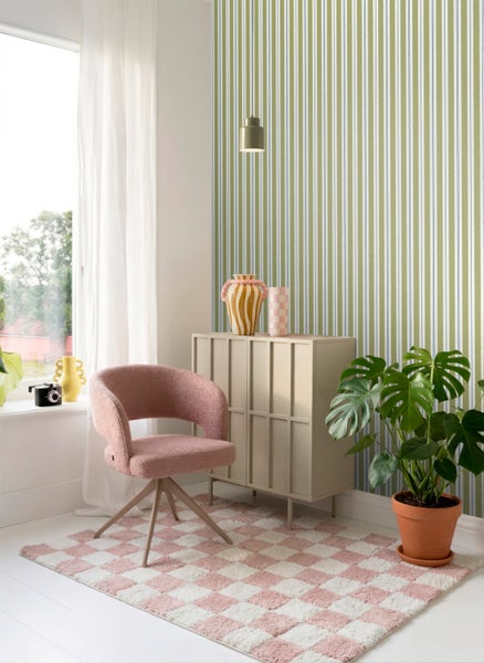 Hell gestalteter Raum mit vertikal gestreifter Tapete, einem rosa Sessel, einem karierten Teppich, einer Kommode und einer Monstera Zimmerpflanze.