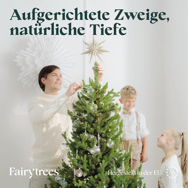 Familie schmückt einen Fairy Trees Weihnachtsbaum mit Sternspitze und durchsichtigen Kugeln in einem hellen Wohnzimmer.