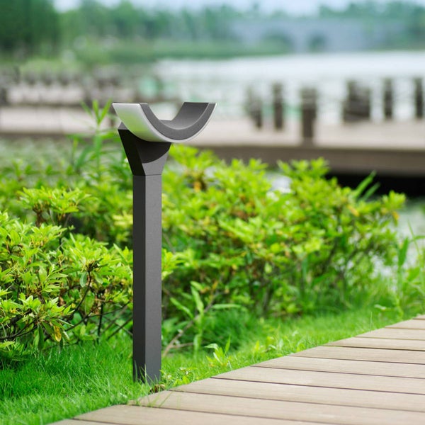 Moderne Außenstandleuchte in Anthrazit mit bogenförmigem Lichtkopf, installiert in einer hellen Gartenanlage neben einem Holzweg.