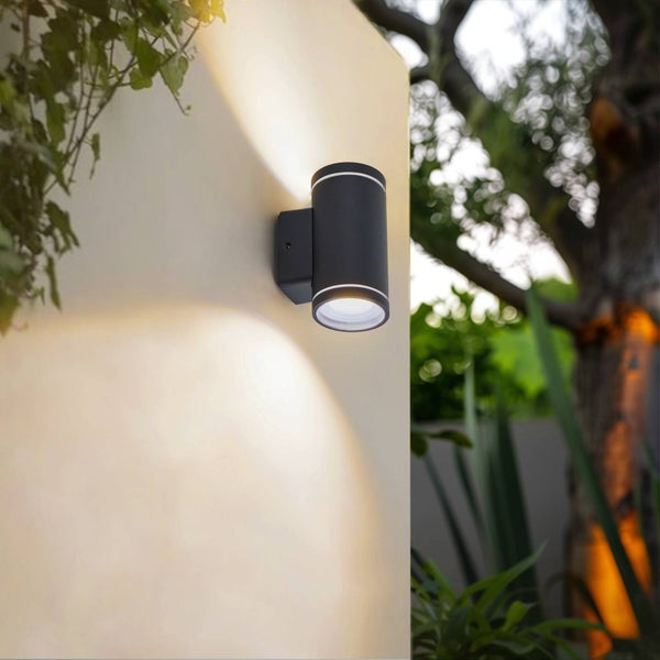 Moderne zylindrische Außenwandleuchte in Schwarz, Up-and-Down-Beleuchtung an einer hellen Gartenmauer.
