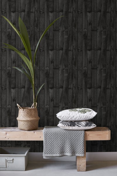 Dekoratives Interieur mit Pflanze, Kissen und Textilien vor einer Wandtapete in Holzoptik.