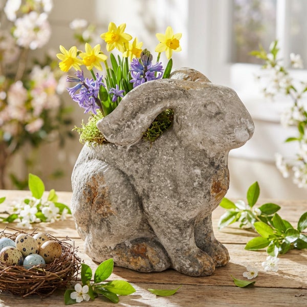 Osterdekoration in Form eines Hasen aus Stein mit Bepflanzung aus Narzissen und Hyazinthen sowie einem Nest mit Eiern auf einem Holztisch.