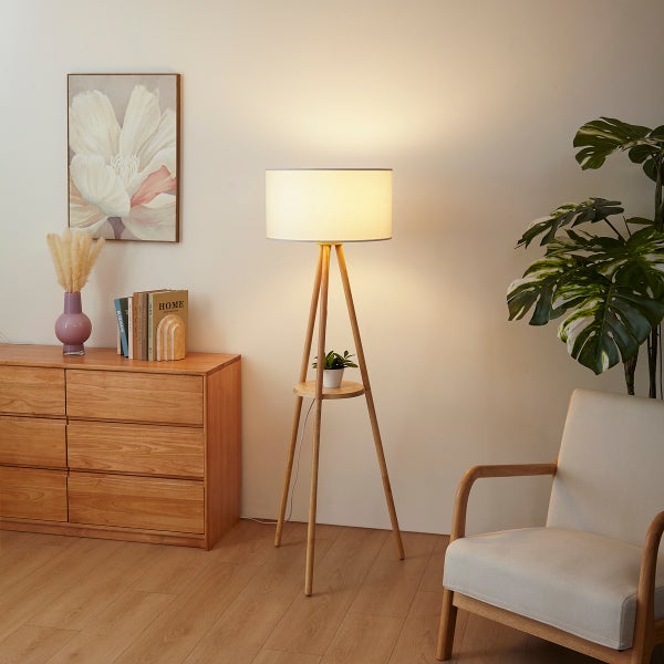 Moderne Dreibein-Stehleuchte aus Holz mit weißem Schirm in einem hellen Wohnzimmer neben einem Sessel und einer Kommode.