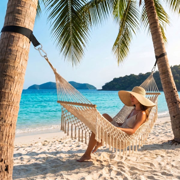 Geflochtene Makramee-Hängematte mit Fransen, befestigt an zwei Palmen an einem Sandstrand, in der eine Frau sitzt.