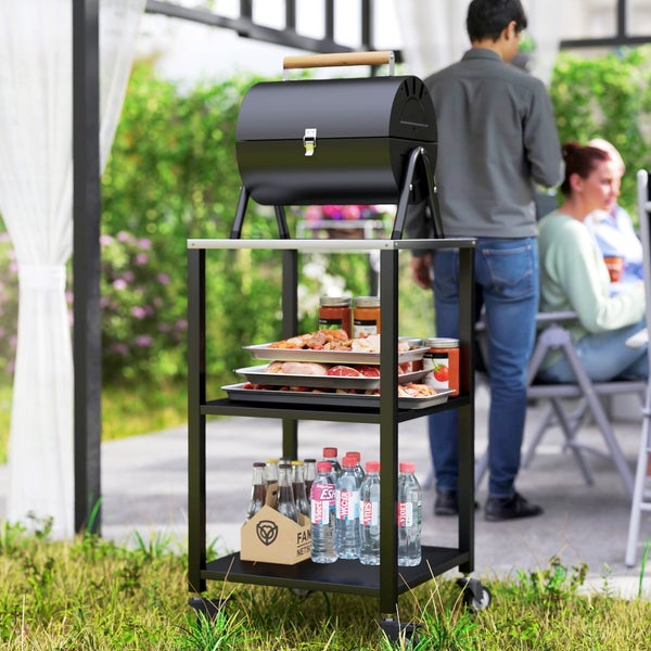 Schwarzer Holzkohle-Fassgrill auf einem Grillwagen aus Metall mit drei Ebenen, Grillgut und Getränken auf einer hellen Terrasse.