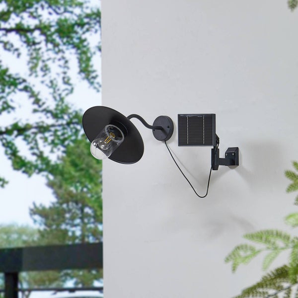 Schwarze Solar-Außenwandleuchte mit Filament-Leuchtmittel und separatem Solarpanel an einer weißen Wand.