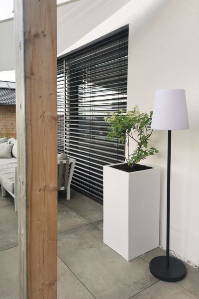 Moderner Außenbereich mit weißem Pflanzkübel, schwarzer Stehleuchte und Außenjalousien auf einer gefliesten Terrasse.