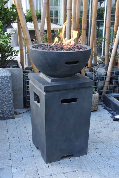 Moderner Gas-Feuertisch mit dunkelgrauem Sockel und Feuerschale mit Lavasteinen und Flammen auf einer Terrasse vor einer Bambuswand.