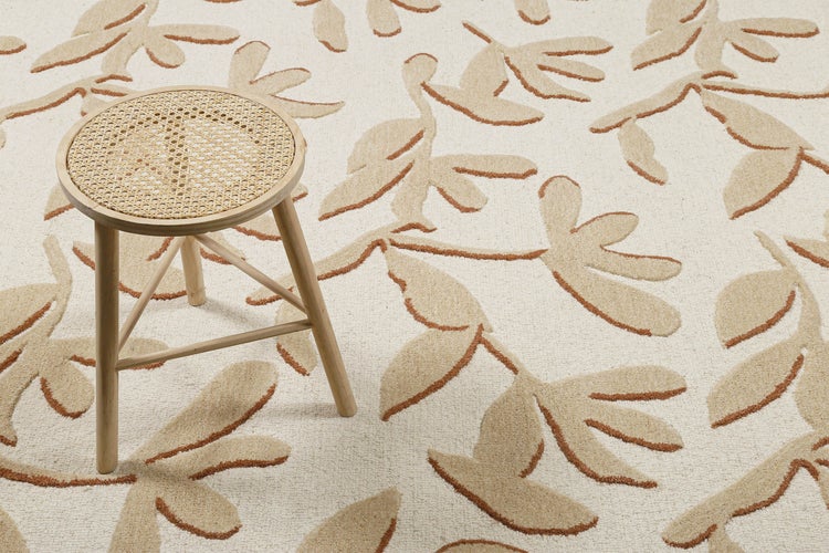 Beige-braun gemusterter Teppich mit Blattmotiven und ein hölzerner Hocker mit runder Sitzfläche aus Geflecht.