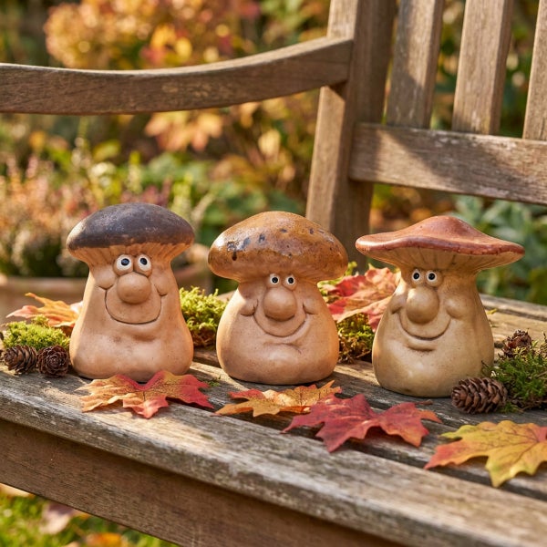 Drei dekorative Pilzfiguren aus Keramik mit freundlichen Gesichtern auf einer Gartenbank mit Herbstblättern und Tannenzapfen dekoriert.