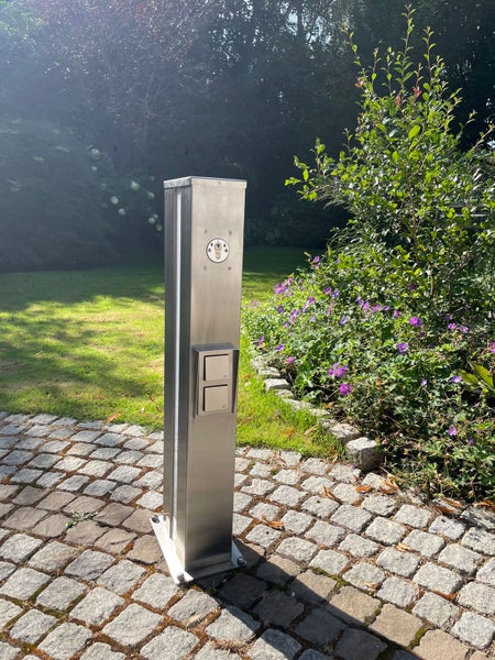 Energiesäule aus Edelstahl mit Außensteckdosen, Lichtleiste und Schlüsselschalter in einem Garten auf Pflastersteinen.