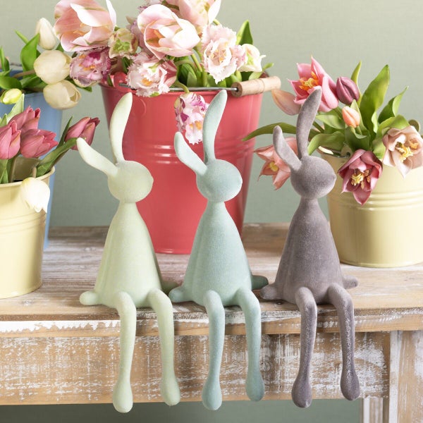Drei sitzende Osterhasen Dekofiguren mit beflockter Oberfläche auf einem Holztisch vor Blumentöpfen mit Tulpen.