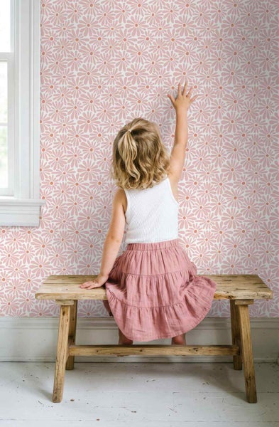 Kind sitzt auf einer Holzbank vor einer Wand mit rosa Gänseblümchen-Mustertapete in einem hellen Zimmer.