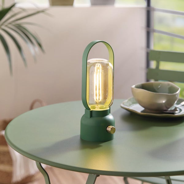 Grüne tragbare LED-Tischleuchte mit Glaszylinder und Tragegriff auf einem grünen Bistrotisch in hellem Außenbereich.
