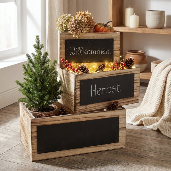 Drei gestapelte Holzkisten mit Kreidetafel zum Beschriften, dekoriert mit einer kleinen Tanne und herbstlichen Elementen wie Kürbis und Zapfen.
