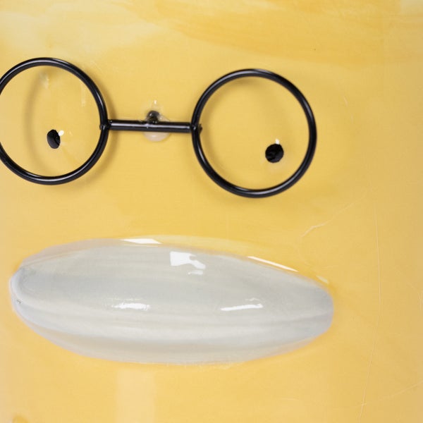 Detailansicht eines gelben Keramikgefäßes mit aufgesetzter Brille und Nase